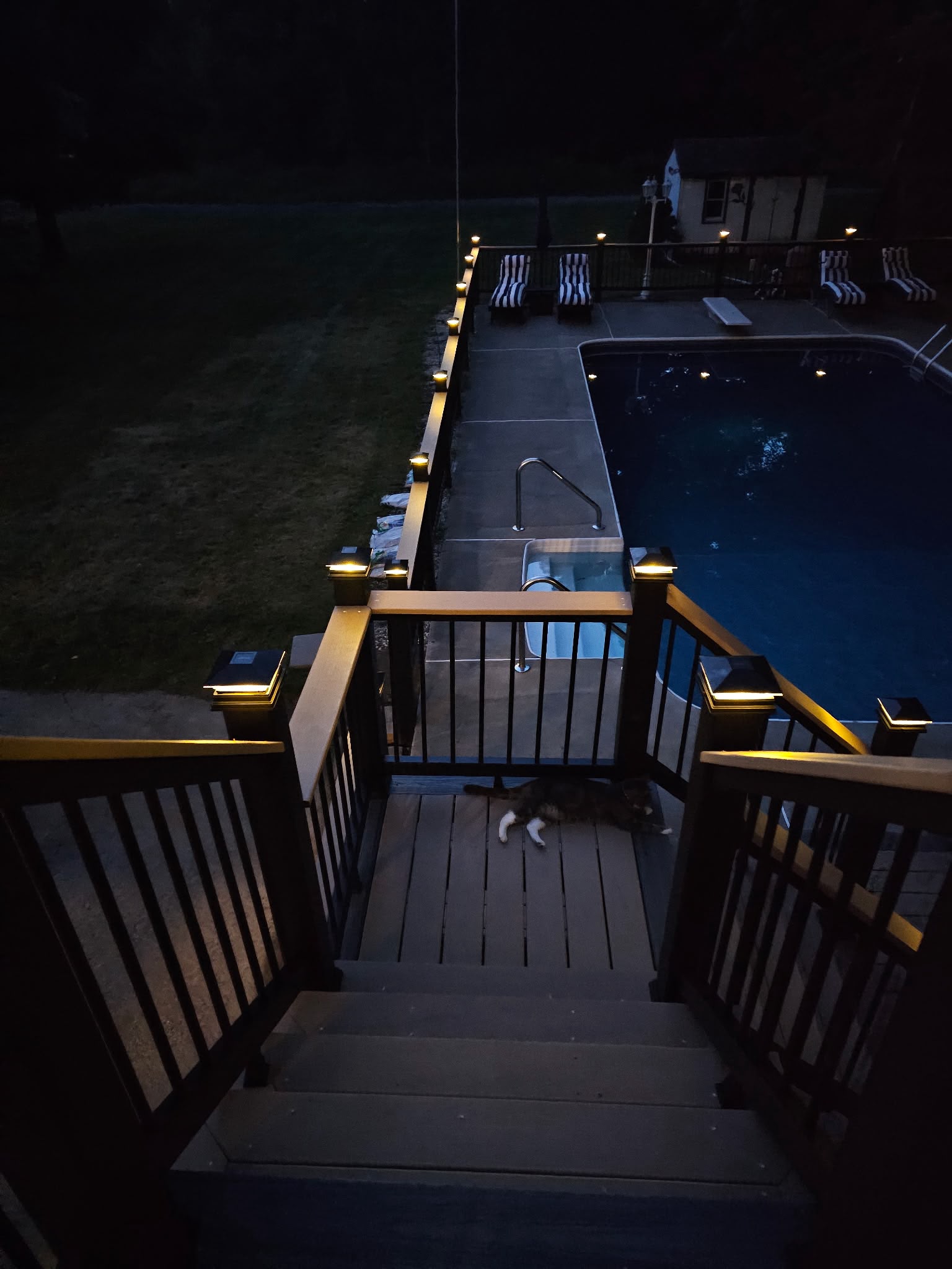 Composite deck stairs leading down to an in-ground pool patio, lit by post cap lights at night