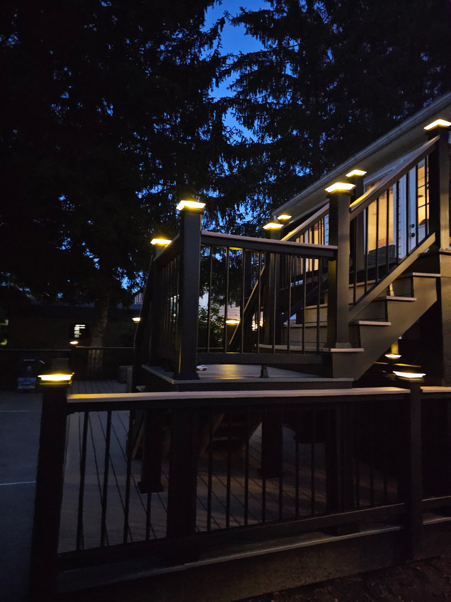 Two-level composite deck with black metal balusters and lit post caps at dusk